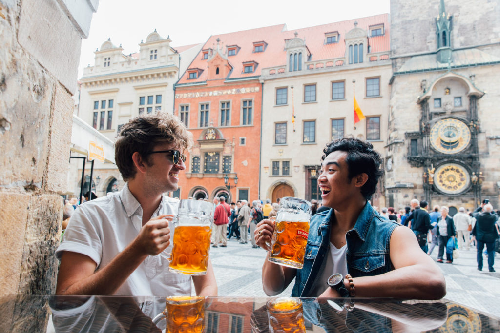 pair of young men sat at a table on a square in Prague holding tankards of beer