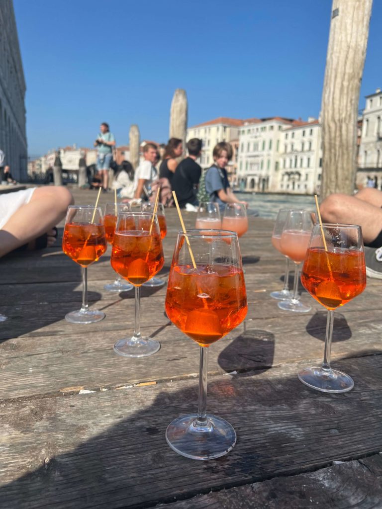 Multiple glasses of Aperol Spritz on decking beside a canal. 