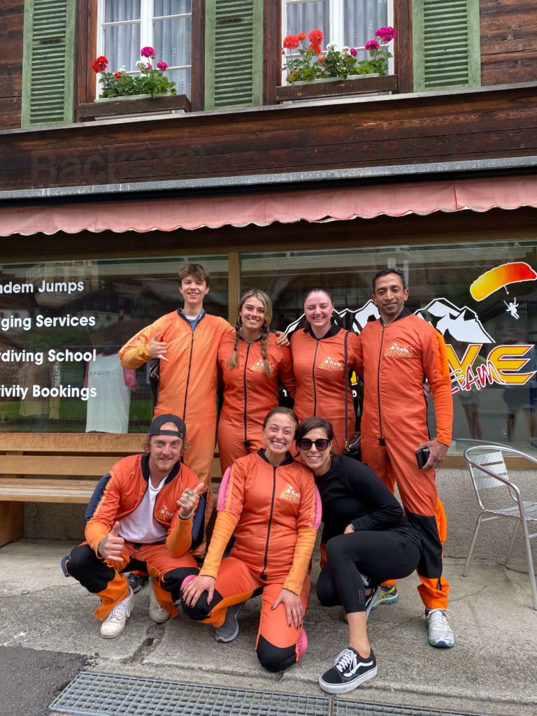Group of travellers dressed in orange jumpsuits, all smiling at the camera in front of skydiving school.