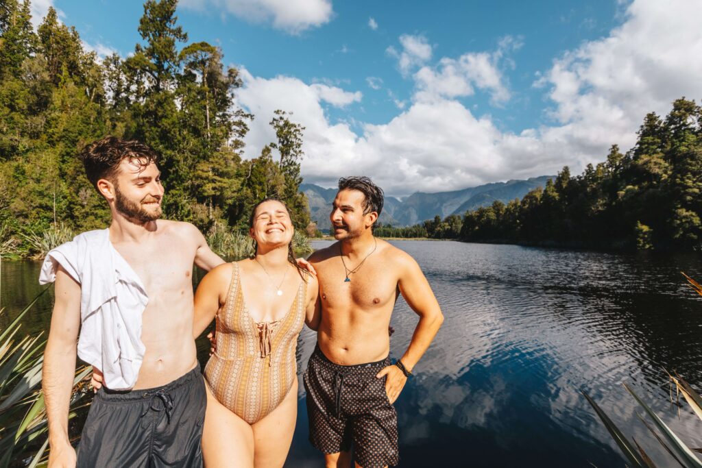 one young female laughing in-between to males, all in swimwear with a calm lake in the background surrounded by trees