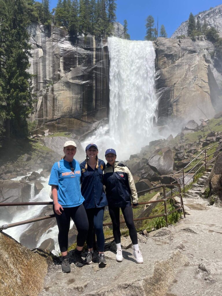 Group of three women wearing caps and activewear smiling at the camera in front of a waterfall at Yosemite National Park.
