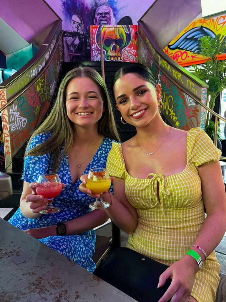 Image of two young women smiling at the camera. One wears a blue dress with small white flowers on it. The other wears a yellow checkered dress. Both are holding cocktails in their right hands.