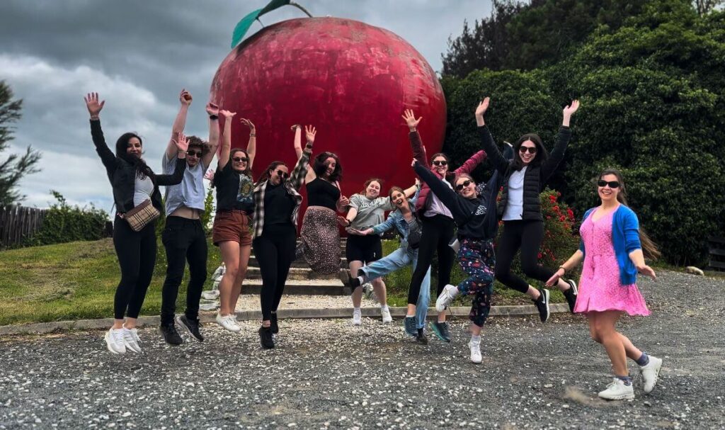 Group of young people in their twenties jumping with their hands in the air in front of a large red apple sculpture.