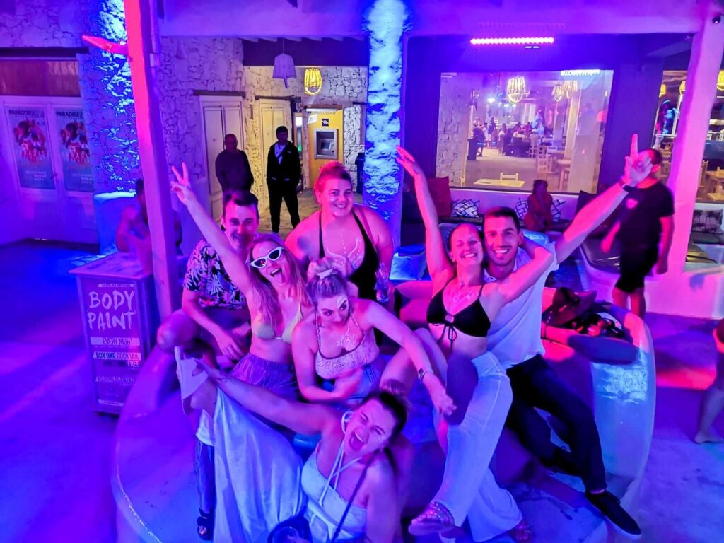 Group shot of 18-29 year old's smiling and raising their hands in Mykonos bar under neon lights
